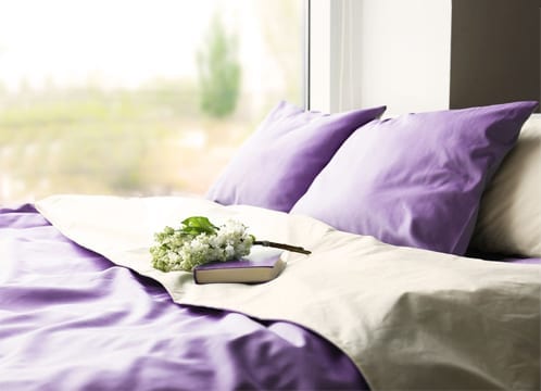 lit soigneusement fait avec des draps et des oreillers violets devant une fenêtre donnant sur un pré vert